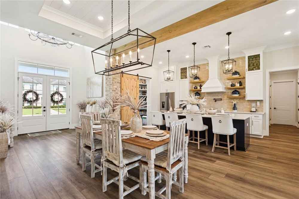 The adjoining dining area is a true showpiece—highlighted by a grand rectangular chandelier, rustic distressed-wood dining set, and decorative natural accents that add texture and warmth. The double French doors flood the space with natural light, opening to a beautiful outdoor view and completing the inviting flow between indoor and outdoor living.
