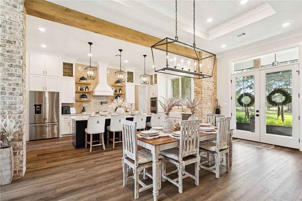 The adjoining dining area continues the warm, organic aesthetic with a distressed wood table and chairs perfectly framed beneath a statement chandelier. Double French doors and transom windows flood the space with natural light, seamlessly connecting indoor comfort with outdoor views. Every element—from the exposed beam detail to the rich wood flooring—creates a timeless blend of sophistication and welcoming warmth, making this the true heart of the home.