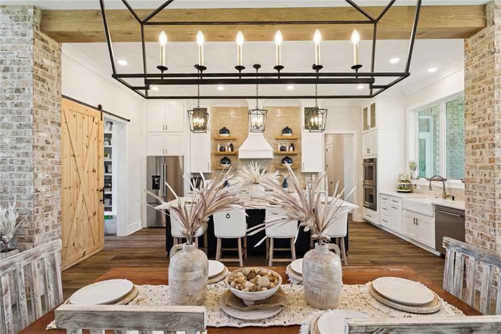 Just steps away, the dining area commands attention with its grand chandelier, exposed wood beam detail, and distressed wood table set for gatherings both intimate and festive. Framed by brick columns and wide-plank flooring, the entire space radiates warmth and cohesion. Beyond the dining area, the living room welcomes you with a cozy fireplace, natural textures, and timeless design elements—making it an ideal space for entertaining or unwinding at the end of the day.