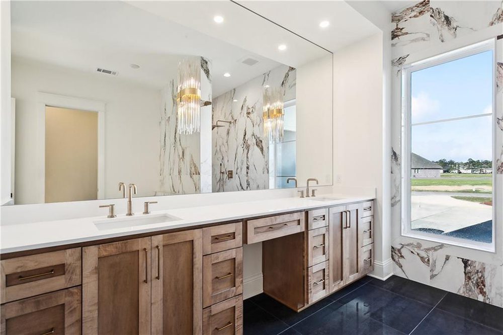 17-Primary Bathroom 1174 Audubon Pkwy A dramatic chandelier with cascading metallic elements introduces a glamorous touch, contrasting beautifully with the earthy cabinetry and modern finishes. The high-gloss black tile flooring grounds the room with a bold, sophisticated base, enhancing the bright and airy ambiance. The open vanity nook is ready for a makeup station or stool, combining function with polished design. This bathroom is a true retreat, balancing opulence and simplicity.