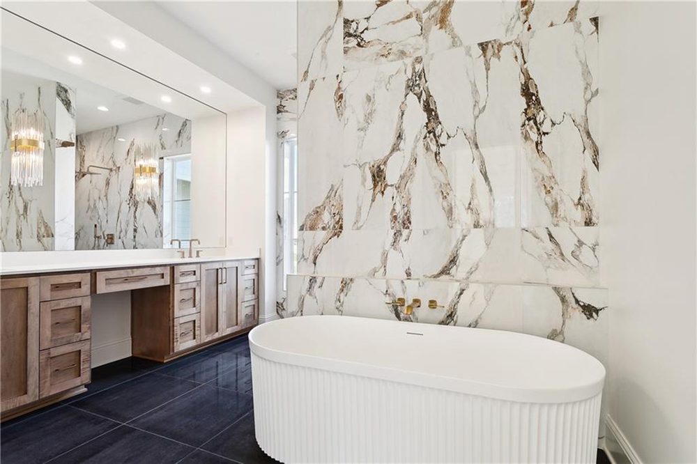 18-Primary Tub 1174 Audubon Pkwy Wall-mounted brass fixtures provide a refined touch, pairing seamlessly with the bath’s minimalist silhouette. The polished black tile flooring adds a striking contrast, grounding the room and enhancing its sense of drama and depth. To the left, the custom double vanity continues the warm- modern theme with natural wood cabinetry, clean white countertops, and integrated sinks. A full-width mirror expands the space visually, reflecting both natural light and the striking chandelier's golden glow. This bathroom embodies high-end, spa-inspired living, blending organic textures and tones with clean, contemporary lines for a space that feels both restful and radiant.