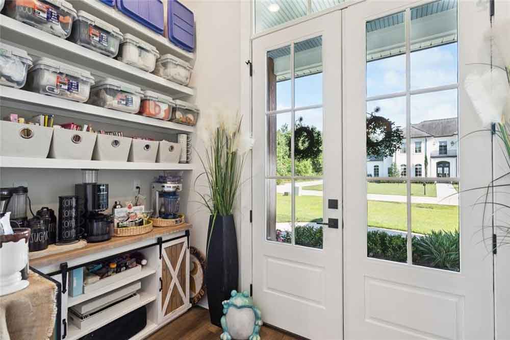 Natural light pours in through the French doors, creating a bright and cheerful atmosphere while offering a glimpse of the landscaped exterior. Every detail has been thoughtfully planned, from the tiered trays and woven baskets to the custom cabinetry that maximizes space without sacrificing aesthetics. This pantry isn’t just for storage—it’s an extension of the home’s warm, inviting personality, blending practical living with farmhouse sophistication.