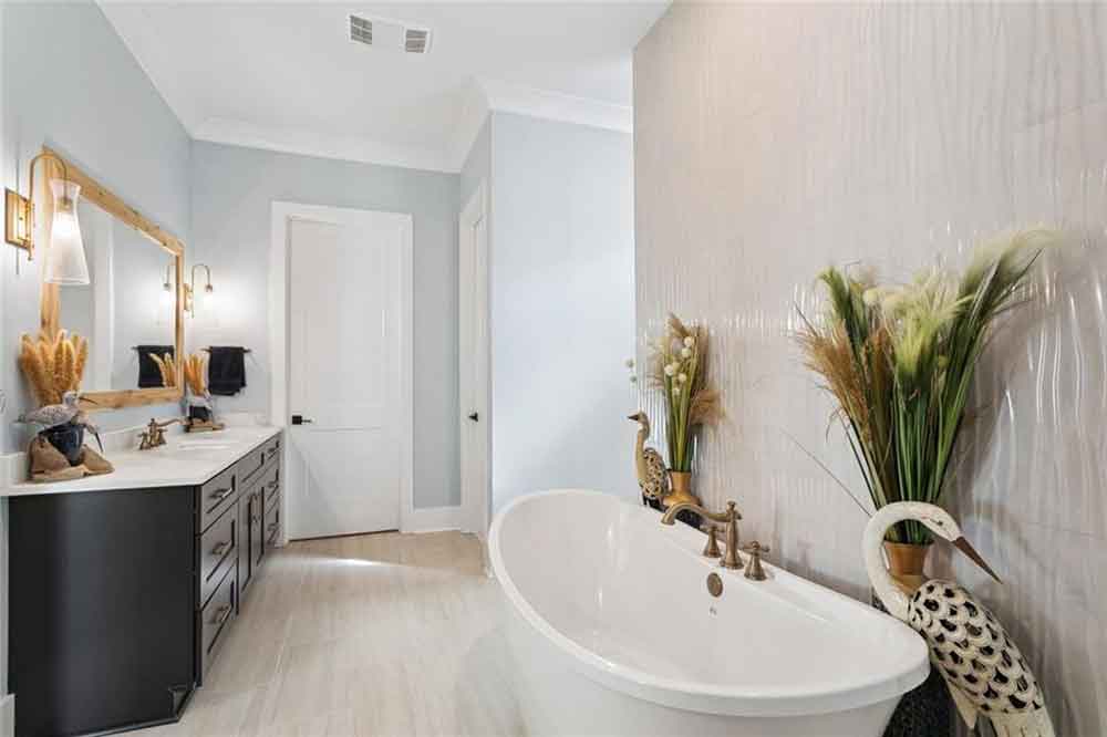 A spacious dual-sink vanity with rich cabinetry and a wood-framed mirror provides both functionality and warmth. Stylish sconces offer ambient lighting, and the light flooring enhances the room’s airy, open feel. Every detail—from the organic greenery to the subtle sheen of the tile—has been thoughtfully curated to create a sanctuary of comfort and elegance.
