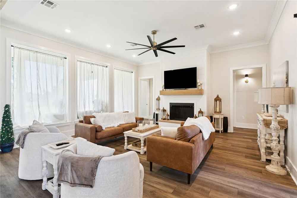 Spacious and sun-filled living room boasting tall windows with sheer curtains, a cozy fireplace with a wood mantel, and a mounted TV as the focal point.