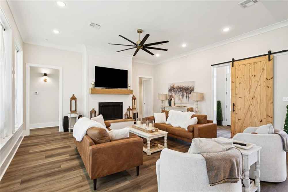 This inviting living room strikes the perfect balance between warmth and modern sophistication. The open-concept design features rich wood-look floors, soft neutral walls, and abundant natural light streaming through multiple large windows. A striking black ceiling fan with sleek blades anchors the space overhead, while the natural wood sliding barn door adds a touch of rustic charm.