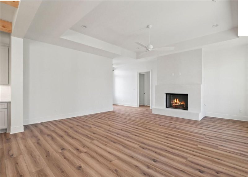 The photo shows a spacious interior with a minimalist design, featuring a white fireplace against a large white wall, light hardwood floors, and a high ceiling with recessed lighting and ceiling fans.